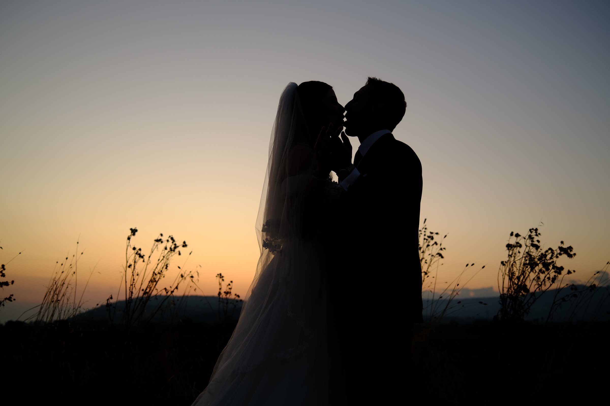 Silhueta dos noivos se beijando contra o céu alaranjado do pôr do sol